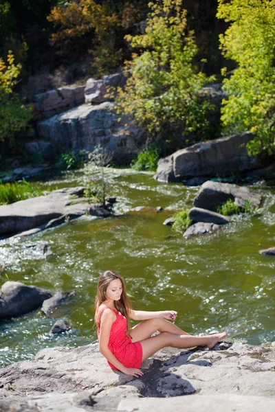Młoda kobieta siedzi na krawędzi urwiska nad szybką górską rzeką latem lub wczesną jesienią na zewnątrz kopia przestrzeń tło — Zdjęcie stockowe