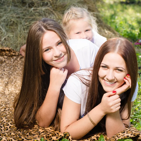 Tres hermanas felices de 3, 13 y 20 años tumbadas en el pajar mirando a la cámara y sonriendo alegremente en el fondo del parque de verano —  Fotos de Stock