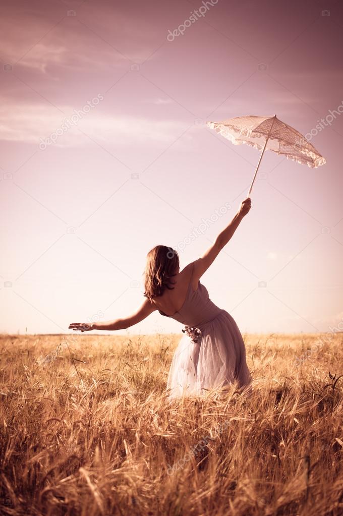 alone in the field: elegant romantic woman in long white dress having fun holding up parasol standing back to camera on blue sky copy space background