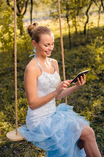 Hermosa mujer joven en vestido de fiesta sentado en columpios y mecanografía en la tableta PC en verde verano u otoño al aire libre copyspace fondo —  Fotos de Stock