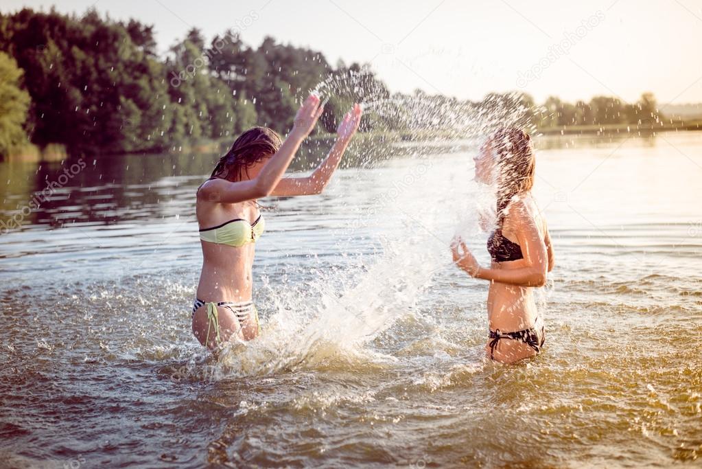 summer fun: filtered image of 2 beautiful young woman or teenage girls best friends having fun and splashing water in river or lake at sunset on sunny outdoors copy space background