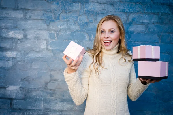 Attraktive blonde junge Frau im Wollpullover, die Geschenke entgegennimmt und glücklich lächelnd auf blauem Backsteinhintergrund lacht — Stockfoto