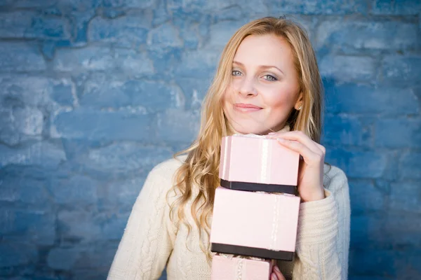 Attraktive blonde junge Frau im Wollpullover, die Geschenke entgegennimmt und glücklich lächelnd auf blauem Backsteinhintergrund lacht — Stockfoto