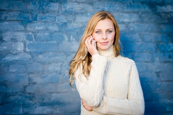 Image de belle jeune femme blonde en chandail de laine blanche parlant sur mobile à fond bleu mur de béton copie espace — Photo