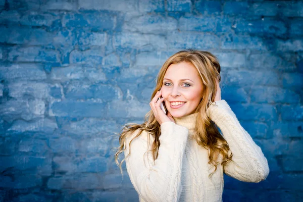 Immagine di bella bionda giovane signora in maglione di lana bianca parlando sul cellulare a parete di cemento blu copia spazio sfondo — Foto Stock