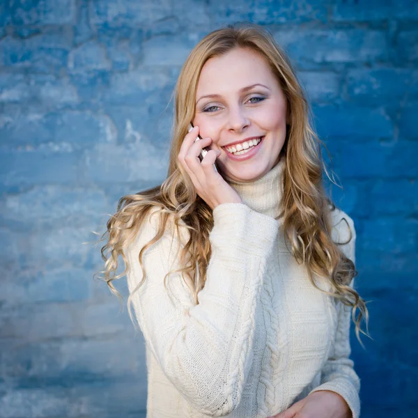 Afbeelding van mooie blonde jonge dame in witte wollen trui spreken op mobiele telefoon op blauwe betonnen wand kopie ruimte achtergrond — Stockfoto