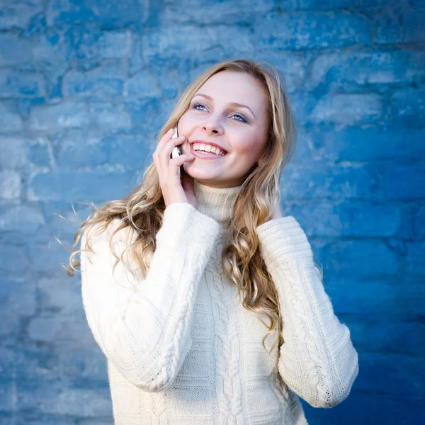 Immagine di bella bionda giovane signora in maglione di lana bianca parlando sul cellulare a parete di cemento blu copia spazio sfondo — Foto Stock