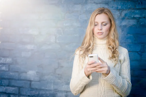 Imagen de la hermosa joven rubia en suéter de lana blanca mensajes de texto mensaje móvil en la pared de hormigón azul copia de fondo de espacio —  Fotos de Stock