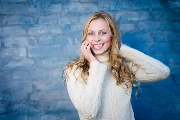 Afbeelding van mooie blonde jonge dame in witte wollen trui spreken op mobiele telefoon op blauwe betonnen wand kopie ruimte achtergrond Stockfoto