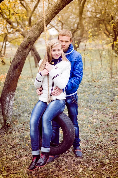 Happy teen couple outdoors on cold autumn day sitting on swings — Stock Photo, Image