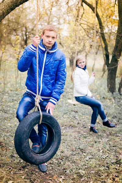 Happy teen couple outdoors on cold autumn day sitting on swings — Stock Photo, Image