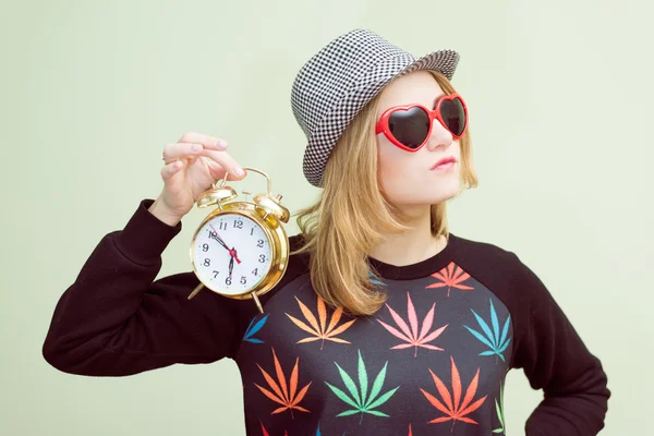 Studio portrait of teenage hipster girl wearing trendy eyeglasses and hat with alarm clock over olive copy space background — Stock Photo, Image