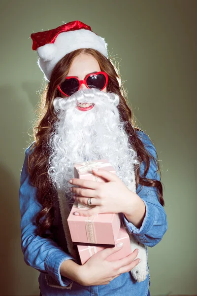 Девушка в рождественской шляпе, очках и бороде Санта держит рождественские шары и подарки — стоковое фото