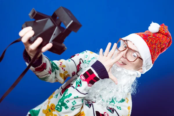 Grappige jonge hipster man in santa baard selfie maken met retro camera op blauwe achtergrond studio portret — Stockfoto