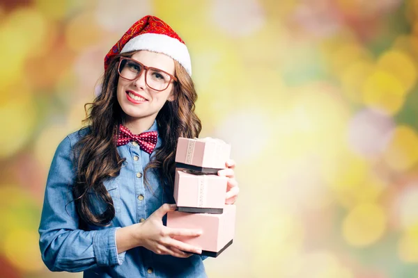 Rolig hipster flicka i super storlek glasögon bär xmas santa hatt anläggningen tre lådor med presenterar under bokeh ljus kopiera utrymme bakgrund — Stockfoto