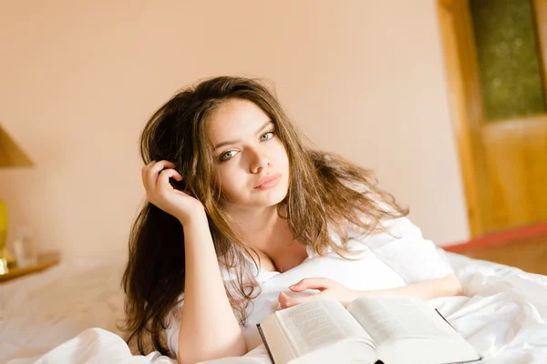 Young lady in white shirt lying on bed reading book — ストック写真