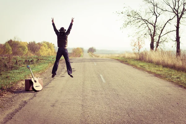 Backview of guitar player in hat jumping on countryside road — Stockfoto