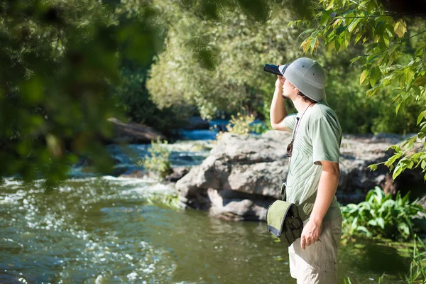 Man in pith helmet looking up through binoculars by  river — Stockfoto