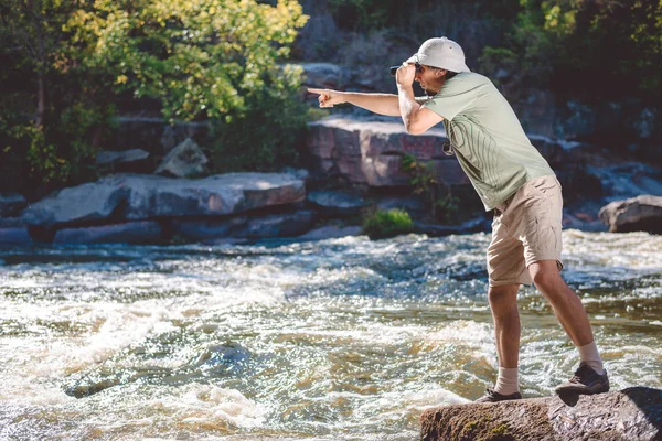 Man in pith helmet standing on rocky riverbank using binoculars — ストック写真