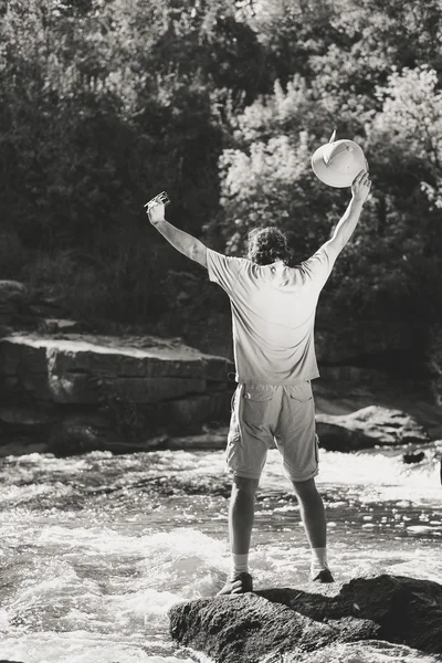 Человек в шлеме стоит на скалистом берегу реки, поднимая оружие. — стоковое фото