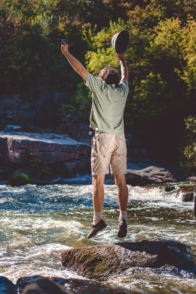 Man holding binoculars jumping up with pithhelmet on rocky riverbank. — Zdjęcie stockowe