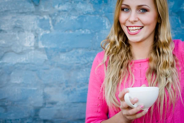 Happy smiling girl holding cup of beverage against brick wall. — Stock Photo, Image