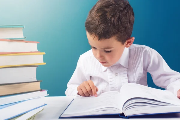 Bonito menino lendo livro difícil na mesa, fundo azul — Fotografia de Stock