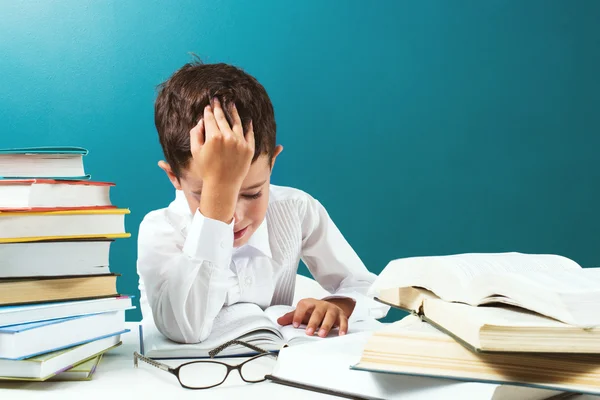 Bonito menino lendo livro difícil na mesa, fundo azul — Fotografia de Stock