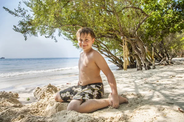 Menino feliz está cavando na areia da praia e construindo edifícios de areia — Fotografia de Stock
