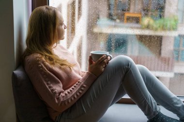 Kadın dinleniyor, yağmurlu bir günde pencere kenarında çay içiyor, rahat, ev, yaşam tarzı, kapalı alan, rahatlama, rahatlama