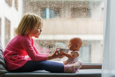 Yağmurlu bir günde elinde oyuncak bir geyikle pencereye oturmuş çay içen küçük bir kız..