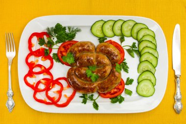 Beyaz tabakta kızarmış domuz sosisi. Sarı masa örtüsü üzerinde taze kesilmiş sebzelerle kompozisyon.