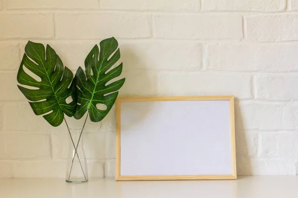 Un lugar de trabajo en casa.Un marco de imagen vacío o una foto y hojas de monstera en un jarrón están sobre la mesa. Estilo escandinavo. —  Fotos de Stock