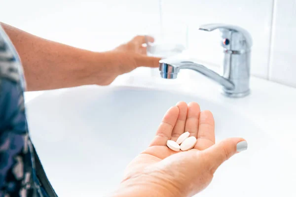 Elderly woman drinks pills or vitamins, the daily dose of medication. Closeup senior Female hands with pills and glass of water at home. Concept of medical treatment, care for the elderly generation