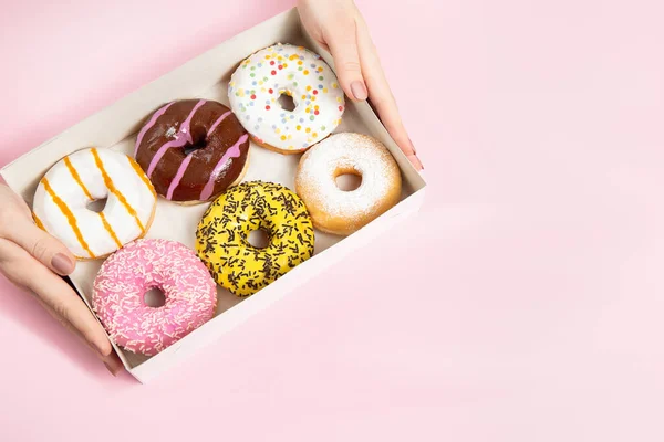 Female hands taking colourful round glazed donuts with sprinkles from donut box on pastel pink background. Different flavors donuts. Concept confectionery store, pastries, coffee shop.