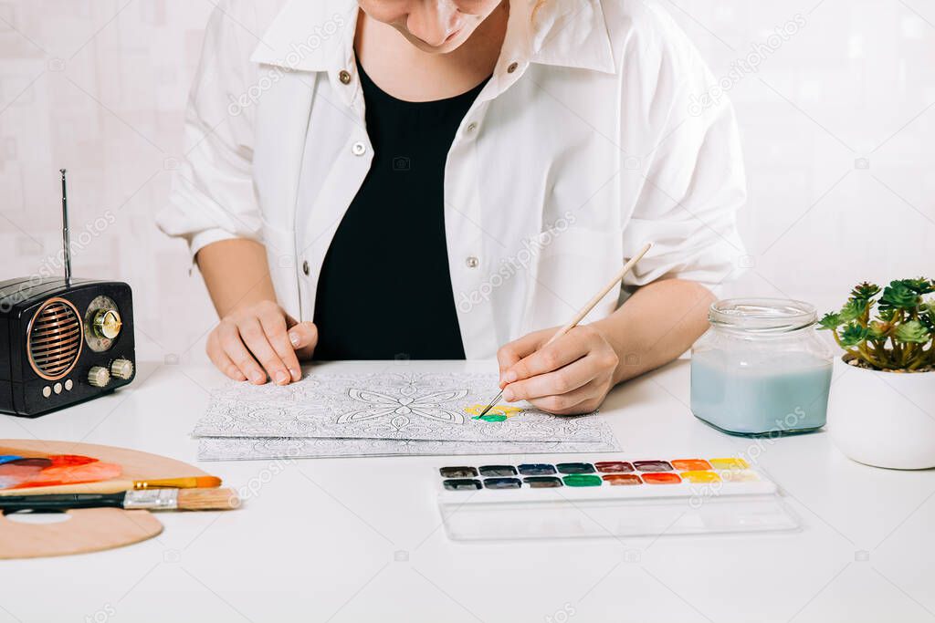 Young woman coloring page antistress at table indoors, mental wellbeing and art therapy. Woman paints a sketch, meditative process of coloring pages.