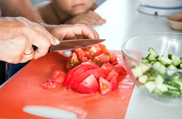 Senior woman grandmother cooking healthy salad for grandson. Grandma cutting tomato by knife on cutting board for dinner