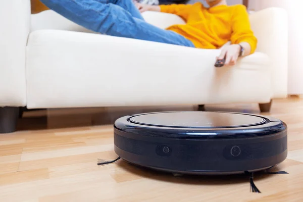 Man resting on the sofa, while the robot vacuum cleaner cleaning living room and dust. Concept time for yourself. Man controlling vacuum cleaner with remote control.