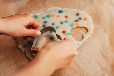 Ellerin yakın çekimi, tatlı hamurdan kalp şeklinde bir kurabiyeyi renkli şeker parçalarıyla kesme çarşafta, Noel ailesi pişirme