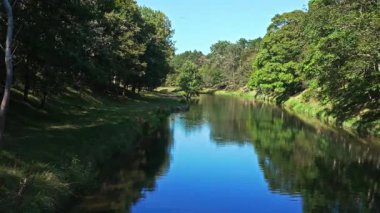 Sakin nehir mavi gökyüzünü yansıtıyor ve yemyeşil ağaçlar yaz güneşinde hafifçe sallanıyor.