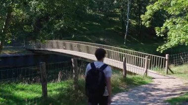 Sırt çantalı bir adam güneş ışığı ve orman yeşiliyle çevrili küçük köprüye doğru yürüyor.