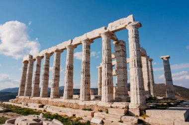 Poseidon Tapınağı 'nın mermer sütunları açık mavi gökyüzüne ve dağlara karşı yükseliyor, antik Yunan mimarisini gösteriyor