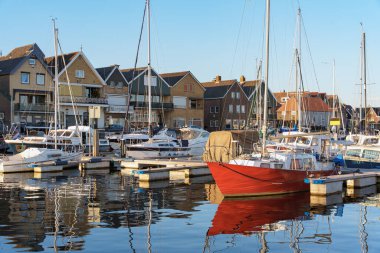 Yat ve teknelerle dolu sakin bir liman parlak sabah gökyüzü altında renkli Hollanda evlerini yansıtır ve hafif su dalgalanmaları, seyahat yaşam tarzı, deniz kenarı mimarisi
