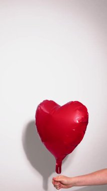 Bir insan eli kırmızı bir kalp şeklindeki folyo balonunu serbest bırakır ve temiz bir duvara karşı dikey bir çerçevede yükselmesini sağlar.