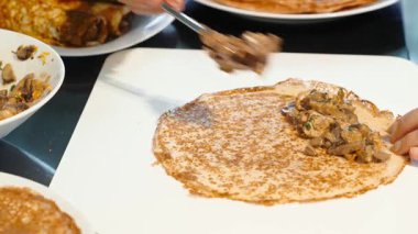 Kişi beyaz tahtada ince ev yapımı krepe sıcak mantar doldurup katlamaya hazırlanıyor. Arkadaki krep yığını görünür..