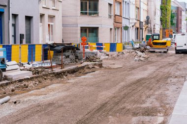 Açık toprak ve ağır makinelerle kazılmış kentsel karayolu güvenlik çitlerinin arkasında devam eden belediye altyapısını gösteriyor