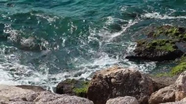 Mavi okyanus dalgaları yuvarlanır ve çarpar, güneş ışığındaki kayalara çarpar, beyaz köpük kıvrılır ve dalgalanır ritimsel deniz kenarında hareket eder, yavaşça uzaklaşır.