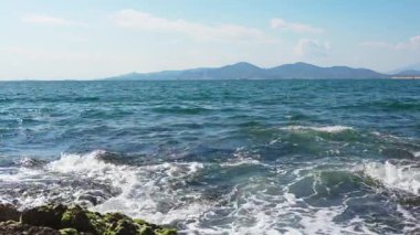 Mavi deniz dalgaları yuvarlanır, dalgalanır ve kayalık kıyıya çarparken köpük uzak dağların yakınındaki açık gökyüzü boyunca sürüklenir.