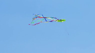 Açık mavi gökyüzünde uçan renkli uçurtma, güneşli bahar gününde rüzgarda savrulan uzun kurdeleler, açık hava aktivitesi, çocukluk eğlencesi.