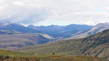 Geniş Yellowstone dağ vadisi manzarası yuvarlanan tepeler ve orman tepeleri dramatik bulutların altında uzanan açık arazi boyunca yayılıyor, sağa dönüyor.